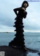 A woman in a black dress standing on a rock by the water.